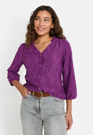 Blusa morada texturizada con escote en V y detalle de botones, mangas largas arremangadas hasta el codo, combinada con jeans de tiro alto en gris claro.