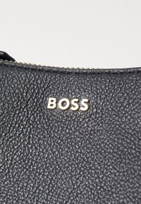 Juodas tekstūruotas odinis maišelis su auksiniu "BOSS" logotipu. Turi užtrauktuką viršuje, pabrėžiantį patvarumą ir meistriškumą.