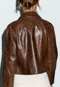 Bruine leren jas met een glanzende afwerking, cropped ontwerp, brede kraag en zichtbare stikseldetails. Achteraanzicht toont een aansluitend silhouet.