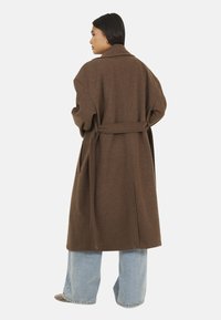 Cappotto oversized marrone con cintura, dotato di un ampio colletto e maniche lunghe. Abbinato a jeans blu larghi. Texture morbida e vestibilità comoda.