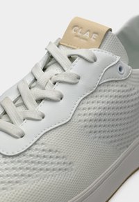 Λευκό αθλητικό παπούτσι με πλεκτή υφή, λευκά κορδόνια, δερμάτινο πάνελ με οπές και μπεζ ετικέτα στη γλώσσα με ανάγλυφο το «CLAE».