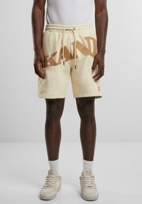 Beige short sweatpants with a textured brown "KAND" logo, elastic waistband, and drawstring. Paired with white socks and cream sneakers.