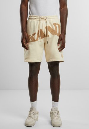 Beige short sweatpants with a textured brown "KAND" logo, elastic waistband, and drawstring. Paired with white socks and cream sneakers.