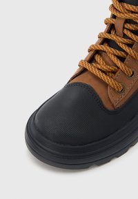 Cizmă de gleznă maro și neagră cu partea superioară texturată, talpă de cauciuc și șireturi multicolore. Are o protecție întărită la vârf și detalii cu ochiuri pentru șireturi.