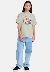 Lysegrøn bomulds t-shirt med et grafisk print, der viser en bjørn og tekst. Sammen med lyseblå vide jeans og mørke sneaks.