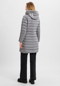 Parka grigia con cappuccio foderato di pelliccia, caratterizzata da sezioni trapuntate orizzontali e un design lungo fino al ginocchio. Indossata con pantaloni larghi neri e stivali robusti.