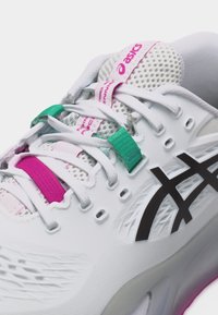 Λευκό αθλητικό παπούτσι ASICS με μαύρο λογότυπο, λευκά κορδόνια και ροζ και τιρκουάζ θηλιές κορδονιών σε λευκό φόντο.