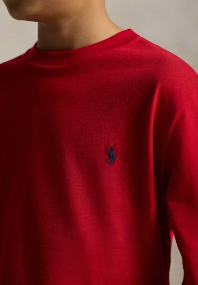 Person wearing a red crewneck sweater with a small embroidered navy blue polo player logo on the left chest.