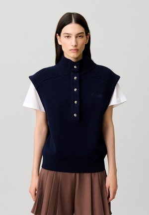 Donna con capelli lunghi e scuri che indossa un gilet in maglia senza maniche blu navy con bottoni sopra una camicia bianca a maniche corte e una gonna plissettata marrone.