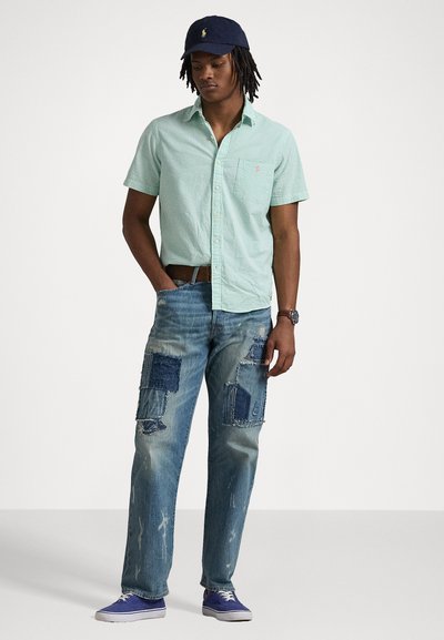 Camicia verde a quadratini a maniche corte, jeans blu strappati con dettagli di toppe, sneakers blu e un cappellino navy con logo.