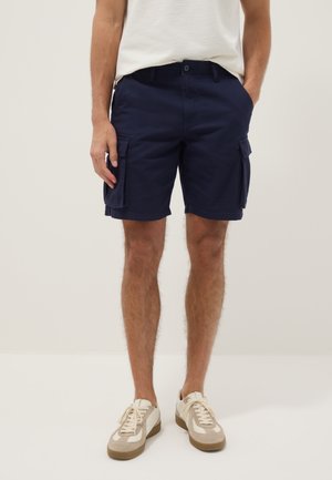 Man met marineblauwe cargoshorts met klepzakken, wit gestructureerd overhemd en beige witte sneakers, staand tegen een neutrale achtergrond.