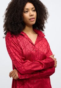 Cămașă lungă cu mâneci lungi din satin roșu, cu un model geometric în formă de inimă, închidere cu nasturi și guler. Textură moale, croială lejeră.