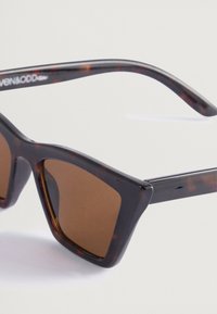 Kattiluukkuja muotoiset tortoise-shell -aurinkolasit ruskeilla linsseillä. Sileä muovikehys, kulmikas muotoilu ja hienovarainen yksityiskohtaisuus templeissa.