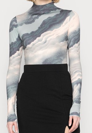 Vrouw draagt een lange mouw coltrui met een grijs-wit wolkenpatroon, ingestopt in een hoog getailleerde zwarte rok met een voorste split.