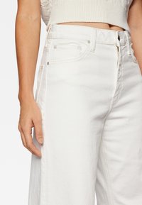 Valkoiset denim-farkut rentolla istuvuudella, joissa on sivutaskut, vetoketjullinen malli ja näkyvät ompeleet. Tasainen pinta ja hillityt yksityiskohdat.