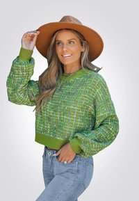 Groene gebreide trui met een getextureerd patroon, geribde manchetten en zoom. Gecombineerd met lichtblauwe jeans en een bruine hoed met brede rand.