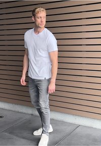 Valkoinen lyhythihainen t-paita, vaaleanharmaat kapeat farkut ja valkoiset lenkkarit. Hahmo seisoo ruskeaa viilutettua seinää vasten.