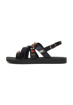 Sandalia plana negra con múltiples tiras, adornada con tachuelas decorativas rojas, verdes y doradas, y un cierre ajustable con hebilla dorada.