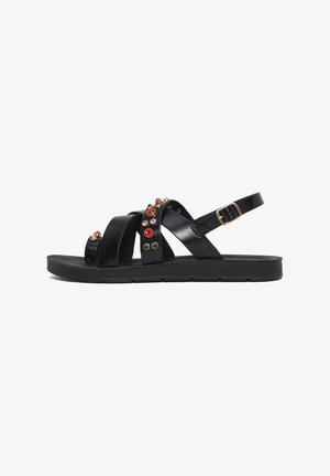 Sandalia plana negra con múltiples tiras, adornada con tachuelas decorativas rojas, verdes y doradas, y un cierre ajustable con hebilla dorada.