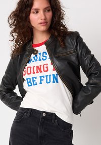 Giacca in pelle nera dal taglio slim, con collo rotondo e tasche con zip. Abbinata a una camicia bianca con scritte rosse e blu e jeans scuri.