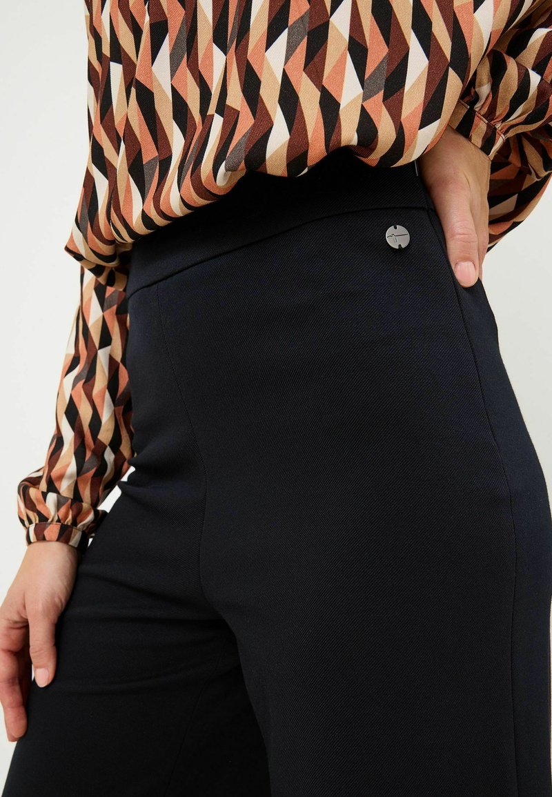 Pantaloni neri con una texture liscia e un design aderente, caratterizzati da un accento in argento con logo in vita, abbinati a una blusa a fantasia.