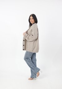 Cárdigan de mezcla de lana beige de corte oversize con seis botones negros, dos bolsillos laterales y puños acanalados. Combinado con jeans de campana azul claro.