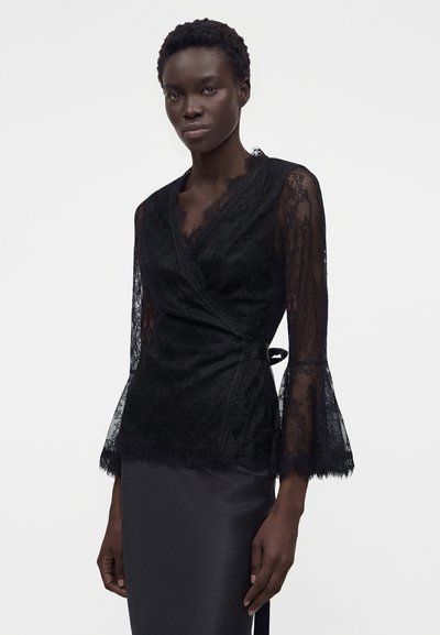 Γυναίκα με εφαρμοστή μαύρη δαντελένια μπλούζα με μανίκια καμπάνα και μαύρη σατέν φούστα, στέκεται μπροστά από έναν απλό λευκό φόντο.