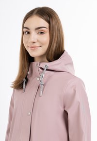 Giacca impermeabile rosa chiaro con cappuccio, dotata di cordini grigi e bottoni automatici metallici. Texture liscia, design casual e senza motivi visibili.