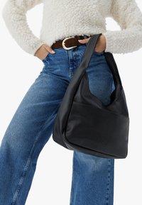 Juodas odinis peties krepšys su laisvu dizainu, lygiu paviršiumi ir įspaustu logotipo detalėmis. Derinamas su šviesiai mėlynomis aukšta juosmeniu džinsomis.