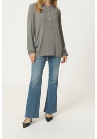 Chemise grise à manches longues avec boutons et design plissé, associée à un jean évasé bleu clair et des escarpins noirs à bouts pointus avec des accents.