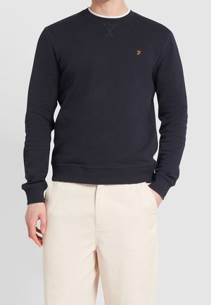 Hombre con sudadera negra de manga larga con un pequeño logo amarillo en el pecho y pantalones beige claros, una mano en el bolsillo.