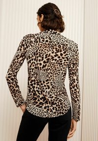 Camicia a maniche lunghe in tessuto beige con un motivo a macchie di leopardo nero e marrone. Texture liscia con orlo curvo sul retro.