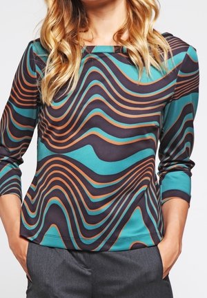 Woman wearing a teal, black, and orange wavy patterned long-sleeve top with three-quarter sleeves and gray pants, hands in pockets.