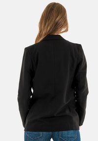 Blazer nero con design su misura, silhouette aderente, rever a punte e texture liscia. Abbinato a jeans in denim blu.