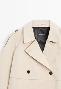 Trench beige con ampio colletto, bottoni neri, dettaglio con bandoliera sulla spalla e fodera interna nera con etichetta Massimo Dutti.