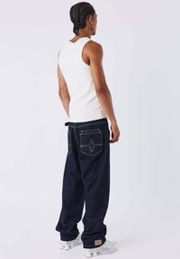 Camiseta sin mangas blanca combinada con jeans de mezclilla oscuros y holgados. Los jeans presentan costuras en contraste y dos bolsillos traseros con un patrón de estrella.