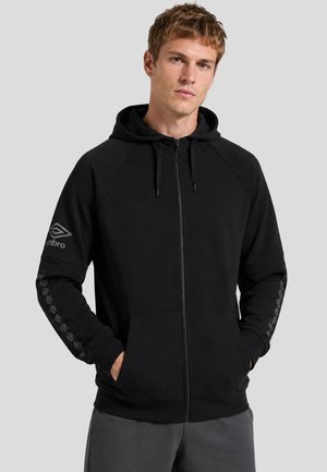 Giovane uomo che indossa una felpa nera con zip e cappuccio e motivi geometrici sulle maniche, in piedi con le mani nelle tasche su sfondo grigio.