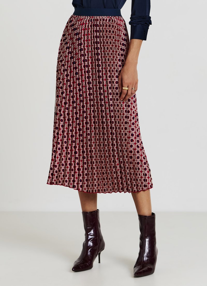 Jupe plissée à motif rouge et crème avec une taille cintrée et un ourlet à hauteur du genou, associée à des bottines brillantes foncées.