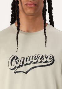 Camiseta de color beige claro con un estampado negro de "Converse", con cuello redondo y mangas cortas. La tela parece suave con un ligero brillo.