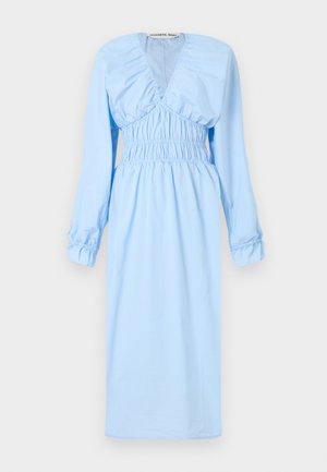 Frau steht in einem hellblauen Midikleid mit langen Ärmeln, geraffter Taille und tiefem V-Ausschnitt, trägt beigefarbene Sandalen mit Blockabsatz auf grauem Boden.