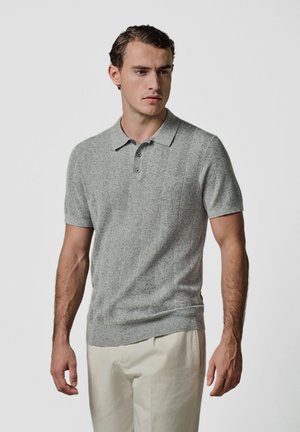 Junger Mann trägt ein graues, kurzärmeliges Poloshirt und helle beige Hose, steht vor einem einfachen hellen Hintergrund.