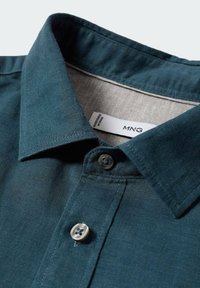 Tumma petrolinvihreä lyhythihainen paita, jossa on levitetty kaulus, kaksi nappia ja kudottu kangasrakenne. Etiketissä lukee "MNG."