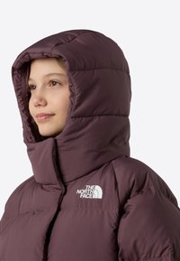 Burgundska puhovka z visokim ovratnikom in veliko kapuco. Zasnovana s prešito strukturo in belim logotipom na prsih. Mehka tekstura.