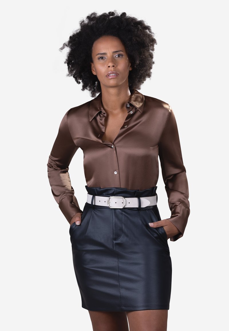 Camisa de satén marrón con botones y cuello, combinada con una falda negra de cuero sintético y un cinturón blanco, que presenta bolsillos y un diseño elegante.