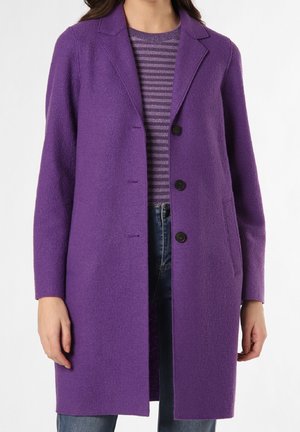 Kvinde iført en midt-lang lilla uldfrakke over en lilla stribet sweater og blå jeans, stående mod en hvid baggrund.