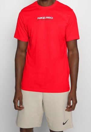 Rode Nike Pro t-shirt met korte mouwen, ronde hals en zwarte Nike-logo tekst op de borst; gedragen met beige shorts.