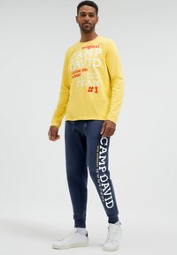 Mann trägt gelbes Langarmshirt und marineblaue Jogginghose mit weißen Turnschuhen, steht vor einem schlichten hellen Hintergrund.