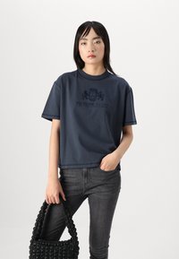 Marineblå T-shirt med korte ærmer, prydet med et logo og tekst i mørkeblå, parret med mørke jeans og en struktureret sort flettet håndtaske.