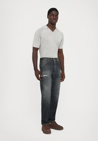 Giovane uomo nero in piedi, che indossa una maglietta a maniche corte di colore grigio chiaro, jeans neri scoloriti con leggere usure e scarpe da ginnastica marroni.