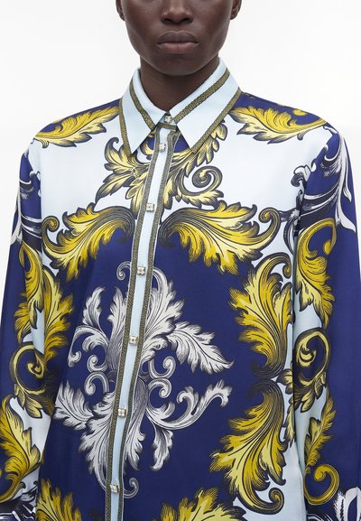 Personne à la peau foncée portant une chemise boutonnée avec un motif floral baroque élaboré en bleu, jaune et blanc et un col pointu.